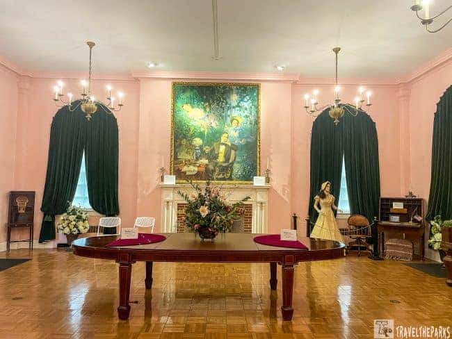  an elegant room in the Stephen Foster Folk Culture Center. The walls are painted a soft pink, contrasting with dark green curtains that frame two windows on either side of a large fireplace mantel. In the center above the fireplace, a large, colorful portrait of a man playing a piano is prominently displayed. The room is lit by chandeliers with multiple bulbs, adding to the classic ambiance. A large wooden table occupies the center of the room, adorned with a floral arrangement. On the table, there are small signs, although the text is not legible. To the right of the fireplace stands a female mannequin dressed in historical attire, adjacent to period furniture, while additional items are positioned on the left of the room.