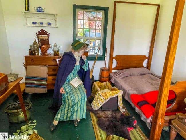 A historically styled room with period furniture and a woman in costume holding a book overlooking a infant's crib.