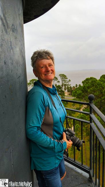 Person smiling on a metal balcony holding a camera, with trees and water in the background at the Crooked River Lighthouse.