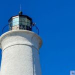 the upper portion of a cylindrical lighthouse located at St. Marks National Wildlife Refuge. The lighthouse is constructed from white bricks, creating a textured appearance. At the top sits a black metal balcony surrounding the lantern room, which has glass panes and a black, conical roof. The sky is a clear, vibrant blue, providing a stark contrast to the white structure. The image captures the lighthouse from a slightly low angle, emphasizing its height and prominence.