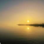 Sunrise over a tranquil Indian River lagoon with sun reflection and faint tree silhouettes.