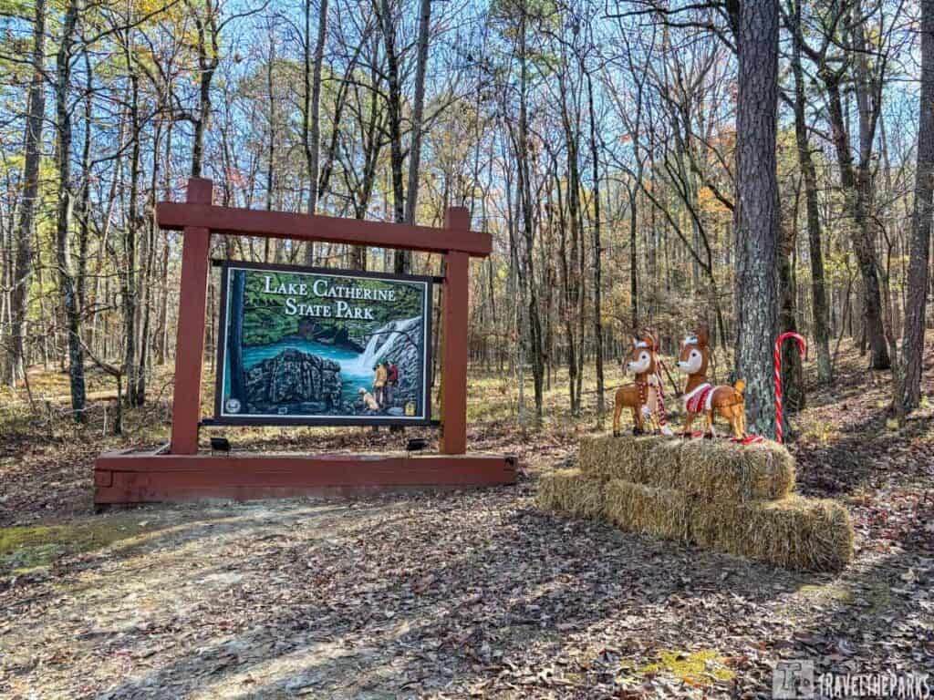 Wooden sign for Lake Catherine State Park with reindeer figurines and a candy cane on hay bales in a forested area.