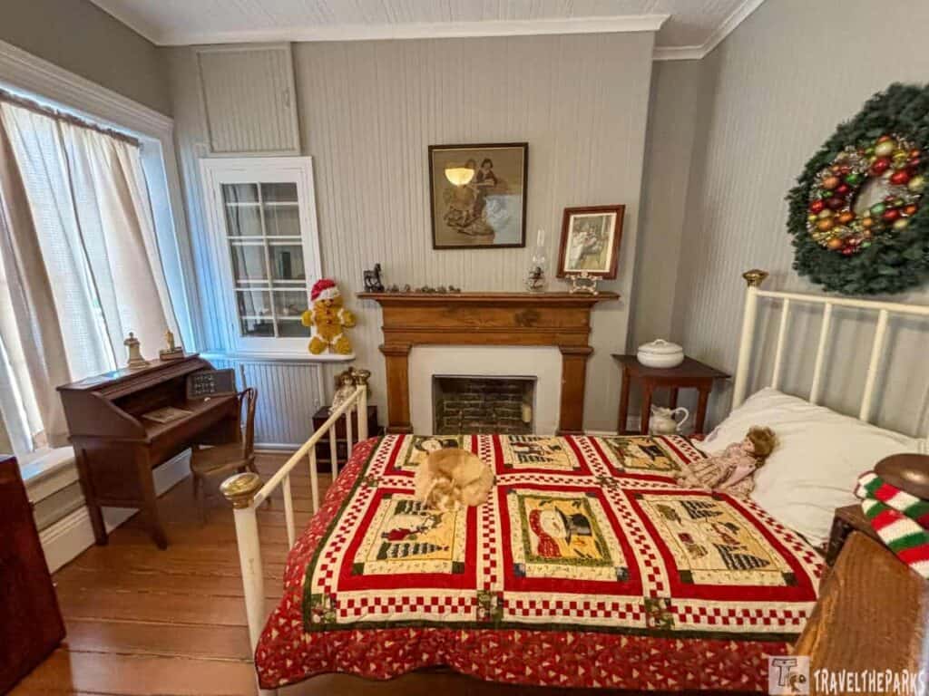 A vintage-style room with a bed, ornate quilt, a mantel with decor, and a desk by a window.