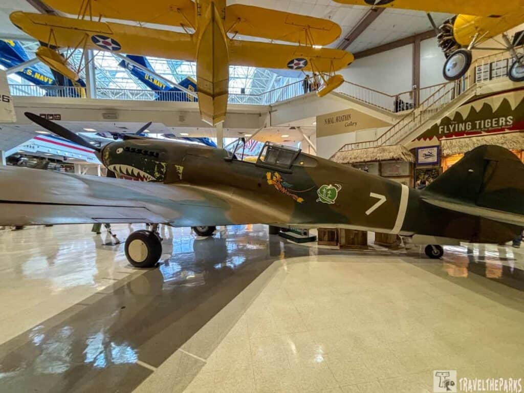 P-40 Warhawk with shark mouth nose art in an aviation gallery, surrounded by other aircraft.