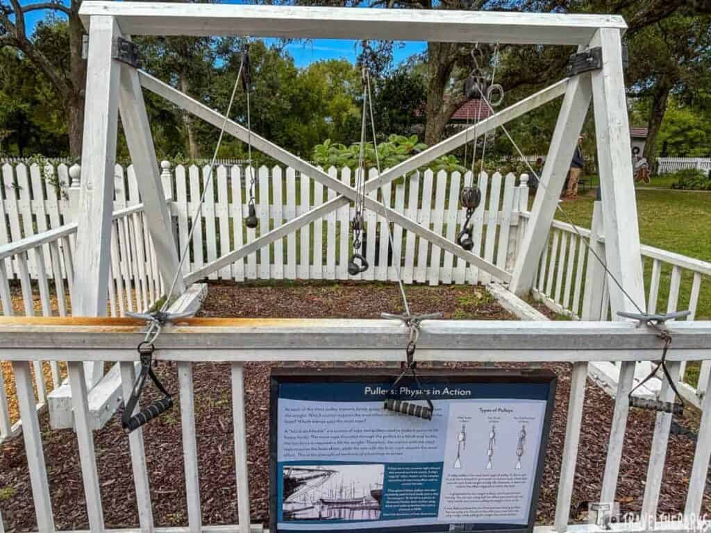 A white wooden A-frame with pulleys and ropes in a garden setting, demonstrating physics principles.