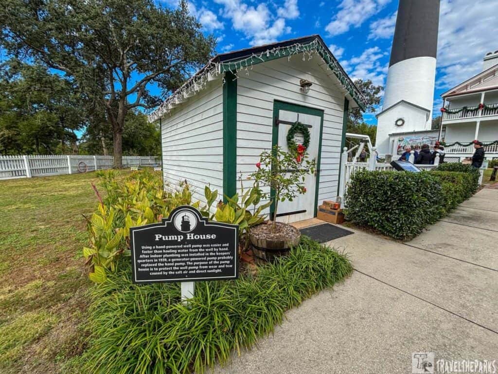 A white wooden pump house with a wreath on the door, accompanied by an informational sign. A lighthouse and a larger building are visible in the background.