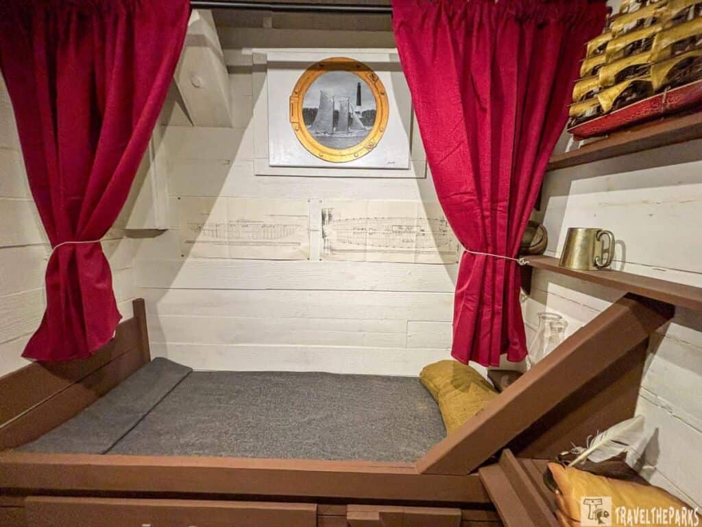 A small room with white walls, a built-in bed with red curtains, and a circular ship image above.