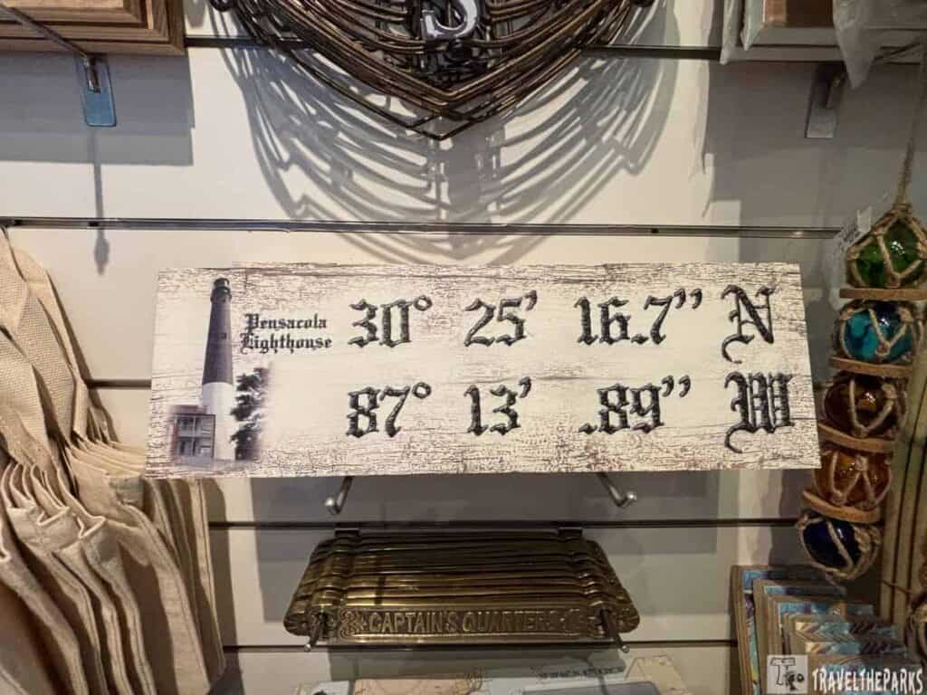 a rustic wooden sign displayed on a shelf. It has a distressed, whitewashed surface and features black, stylized text and an image of a lighthouse. The left side of the sign has a vertical image of a tall lighthouse with trees beside it. The right side displays coordinates in large, ornate black letters. Surrounding the sign, there are various items including what appear to be fabric bags on the left, a metal item below labeled "CAPTAIN'S QUARTERS," and colorful glass buoys on the right.
