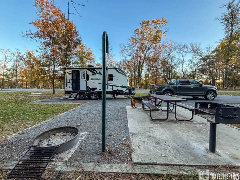 Campsite with trailer and truck at Moro Bay State Park, featuring a picnic table, fire pit, and autumn trees.
