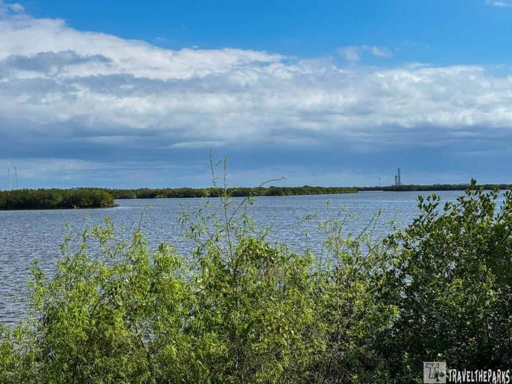 A calm body of water bordered by green foliage and under a partly cloudy blue sky with a tall structure in the distance.