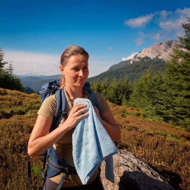 A person in hiking gear holding a blue towel, standing in a mountainous landscape with trees and blue sky.