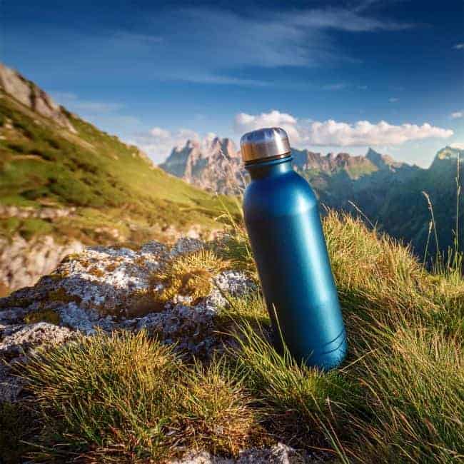 A blue metallic water bottle on grass with mountains in the background under a clear sky.