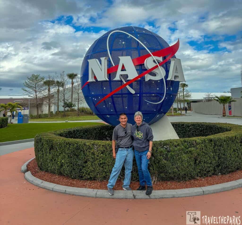 Two people standing in front of the NASA globe logo with a blue sky and clouds in the background.