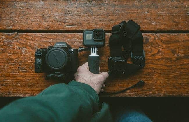 Hand holding a small camera with a grip on a wooden surface, next to a larger camera and a head strap with a camera.