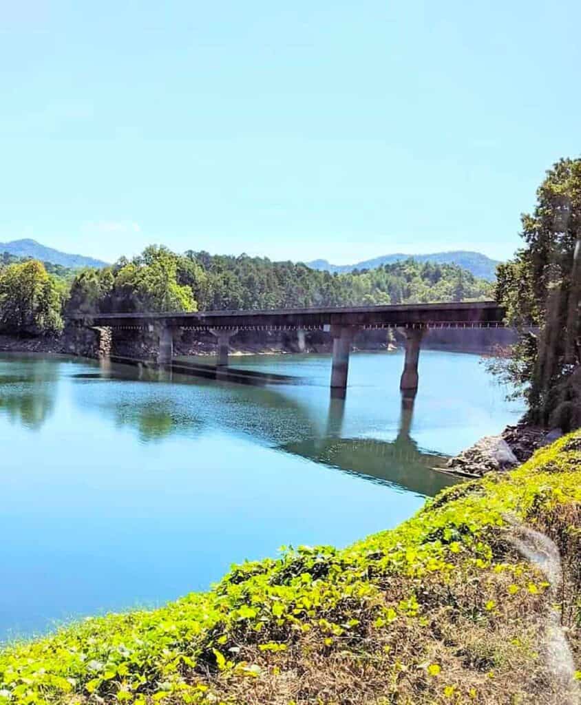 A railway bridge crosses a calm Fontana Lake surrounded by green trees and hills under a clear blue sky.