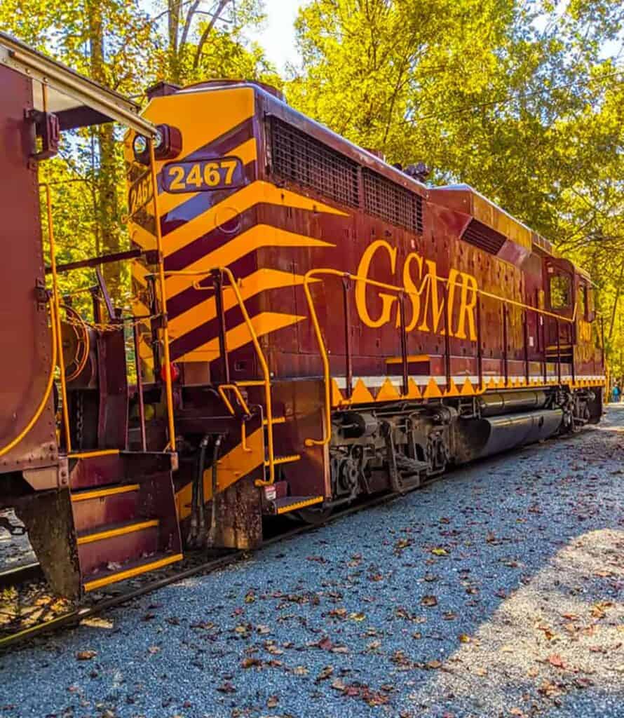 A maroon , Great Smoky Mountains Rail train engine parked on a gravel track surrounded by trees.