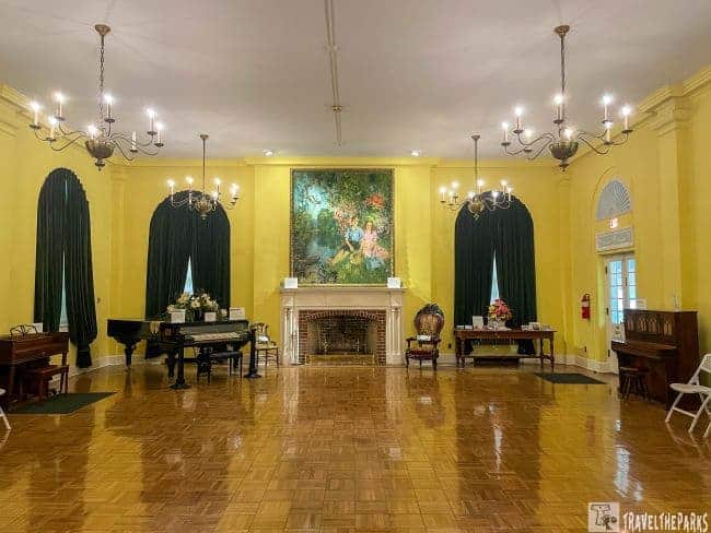 Elegant room with yellow walls, a large painting above the fireplace, pianos, chandeliers, and green drapes.
