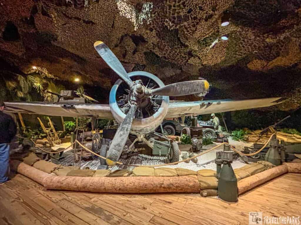 Indoor display of a vintage propeller aircraft with a military-themed setting, including sandbags, camouflage netting, and mannequins.