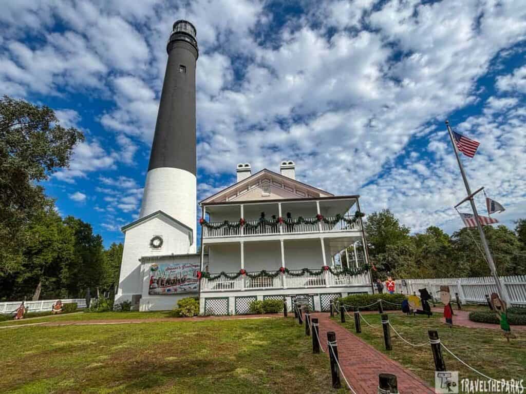 Pensacola lighthouse with a black top and white base beside a decorated two-story building under a bright sky.