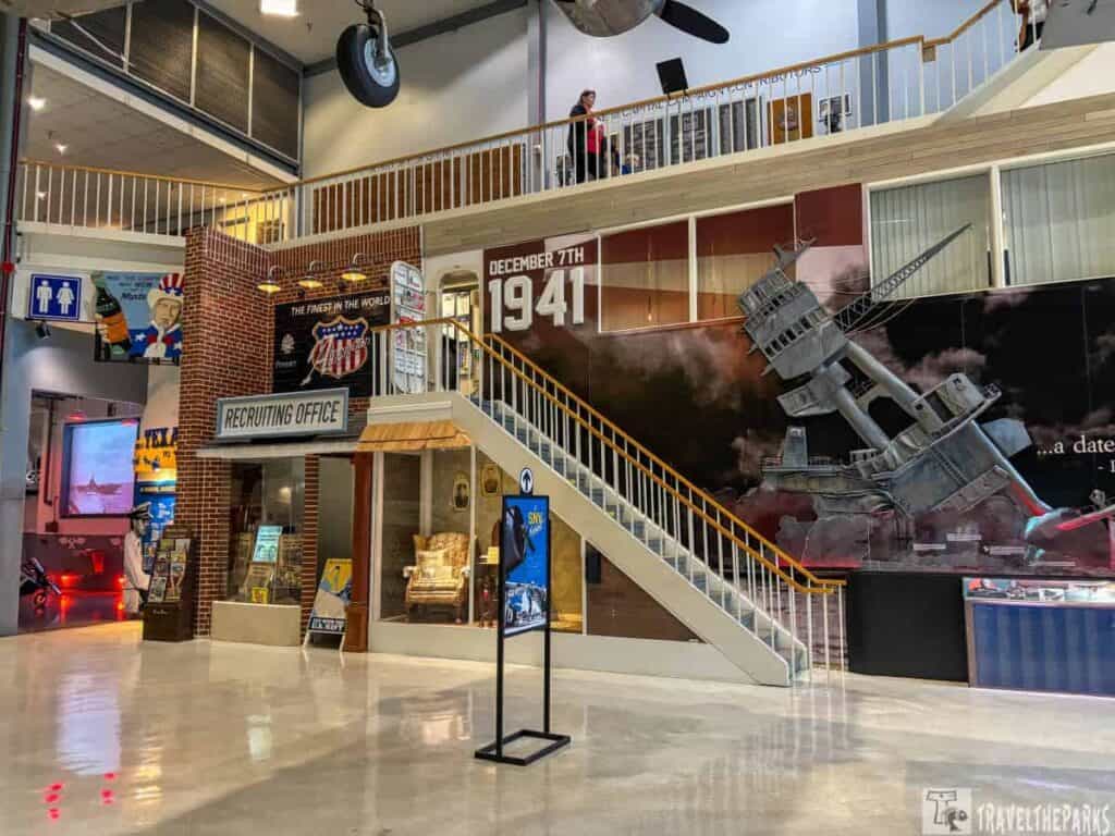 Interior of a World War II-themed museum exhibit with displays, a recruiting office, a staircase, and a mural of a battleship with "December 7th, 1941."