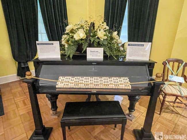 A dark pianola with floral arrangement and informational plaques in a room with yellow walls and green curtains.