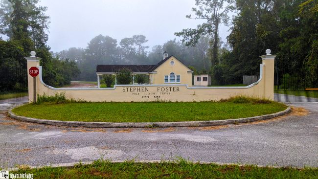 Entrance to Stephen Foster Folk Culture Center State Park with a light yellow wall and building framed by trees.