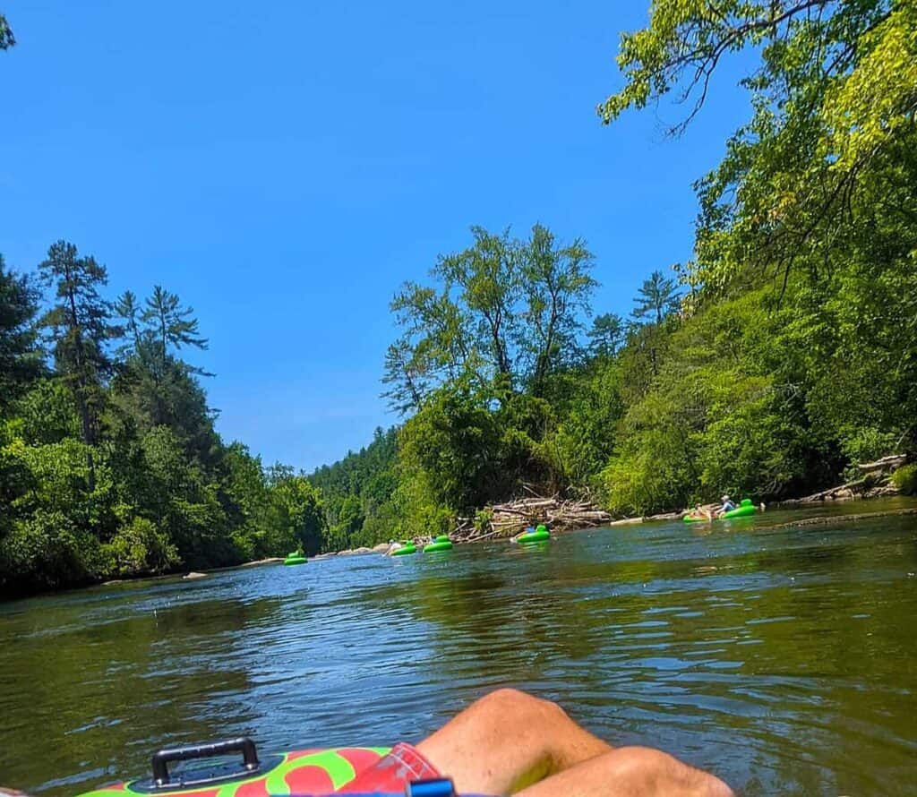 Person floating on a river in an inflatable tube with others in the distance, surrounded by lush greenery and under a clear blue sky.