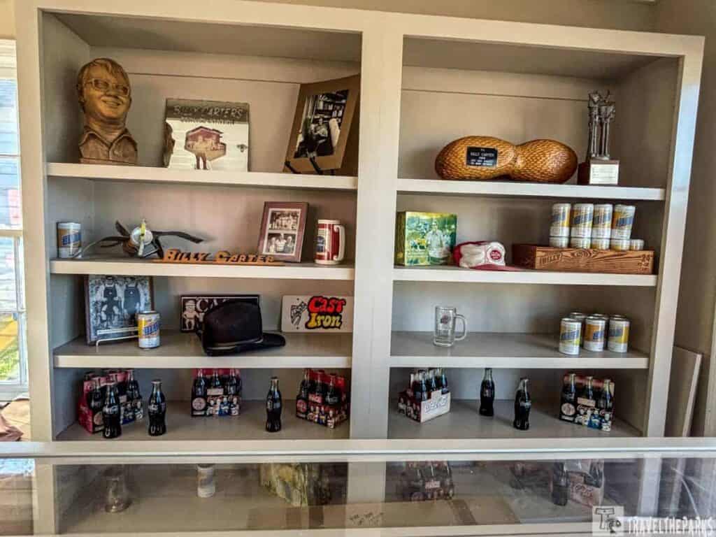 Display shelf with memorabilia including a bust, photos, model airplane, beer cans, soda bottles, a peanut sculpture, and a trophy.