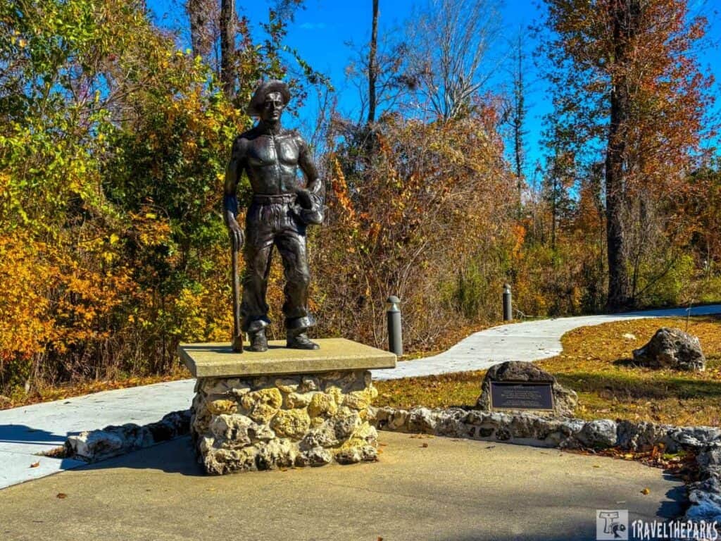 Bronze CCC Worker statue of a man in a natural park setting with autumn trees and a paved path. Floirda Caverns State Park.