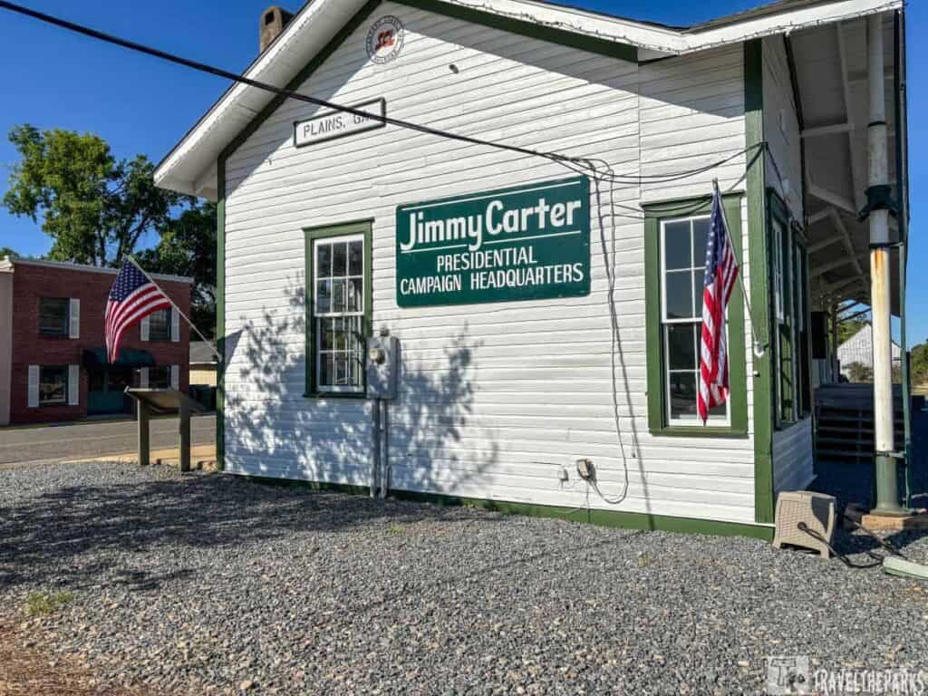 White wooden building with green trim labeled "Jimmy Carter Presidential Campaign Headquarters," with American flags and gravel foreground.