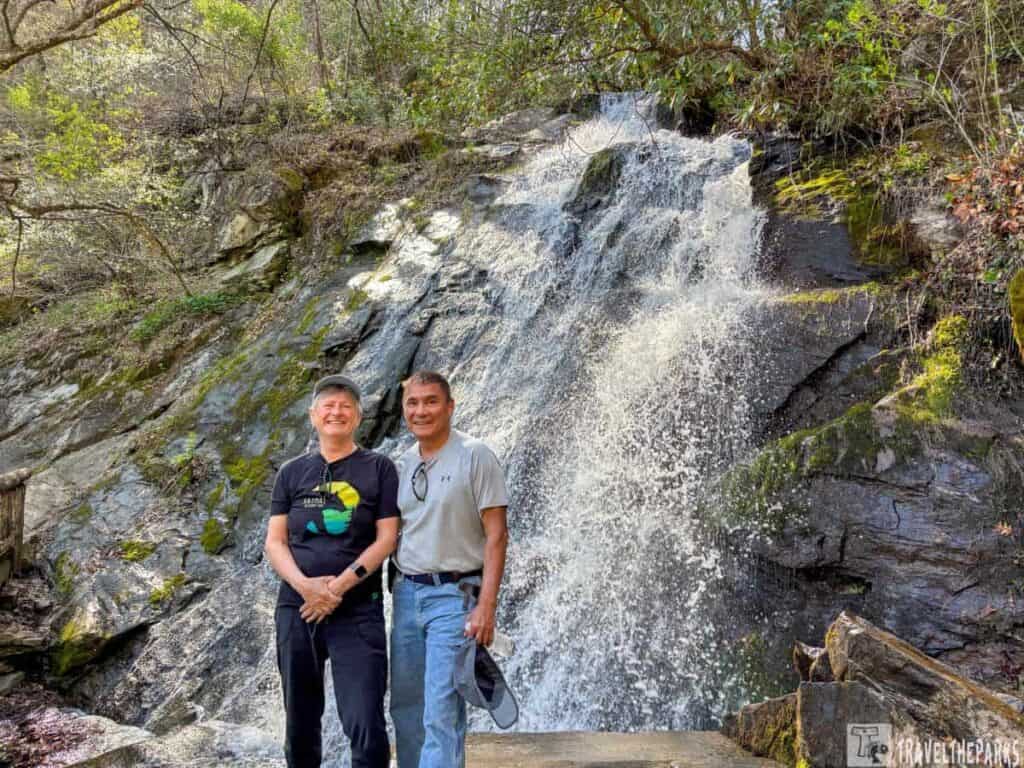 Two people smiling in front of a waterfall on the Juney Whank Falls Trail Loop with surrounding greenery.