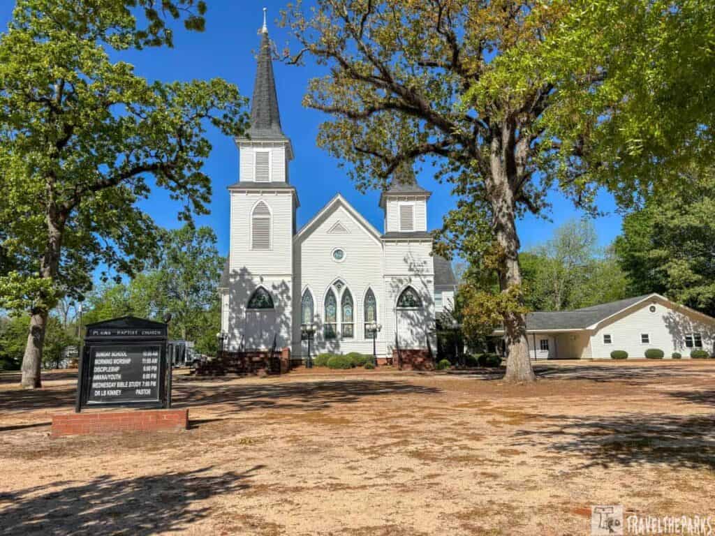 A white wooden church with a tall spire and stained glass windows, surrounded by trees. A sign with service times is in the foreground.