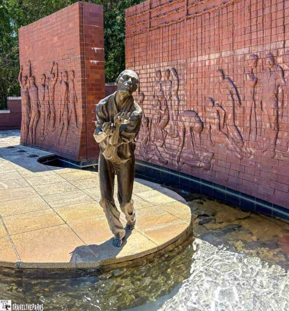 Bronze statue of a man in front of a brick relief mural depicting multiple figures at Andersonville The Price of Freedom Paid