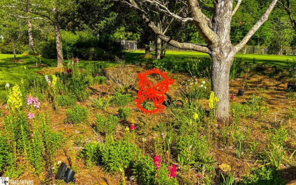 Roslynn Carter Butterfly trail garden with a red butterfly decorative sign, colorful flowers, and a large tree.