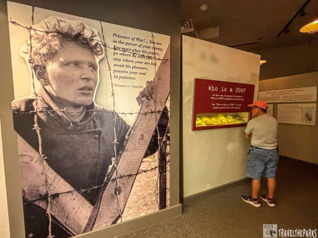 a museum exhibit at the Andersonville National Historic Site. On the left side, there's a large black-and-white photograph of a man in uniform holding onto barbed wire. This photograph is part of a display with a quote from Winston S. Churchill about prisoners of war. The right side features a section with a red panel titled “Who is a POW?” It is mounted above a display case containing several illuminated items. A person wearing a coral-colored cap, a gray shirt, blue shorts, and sneakers is closely viewing the display.