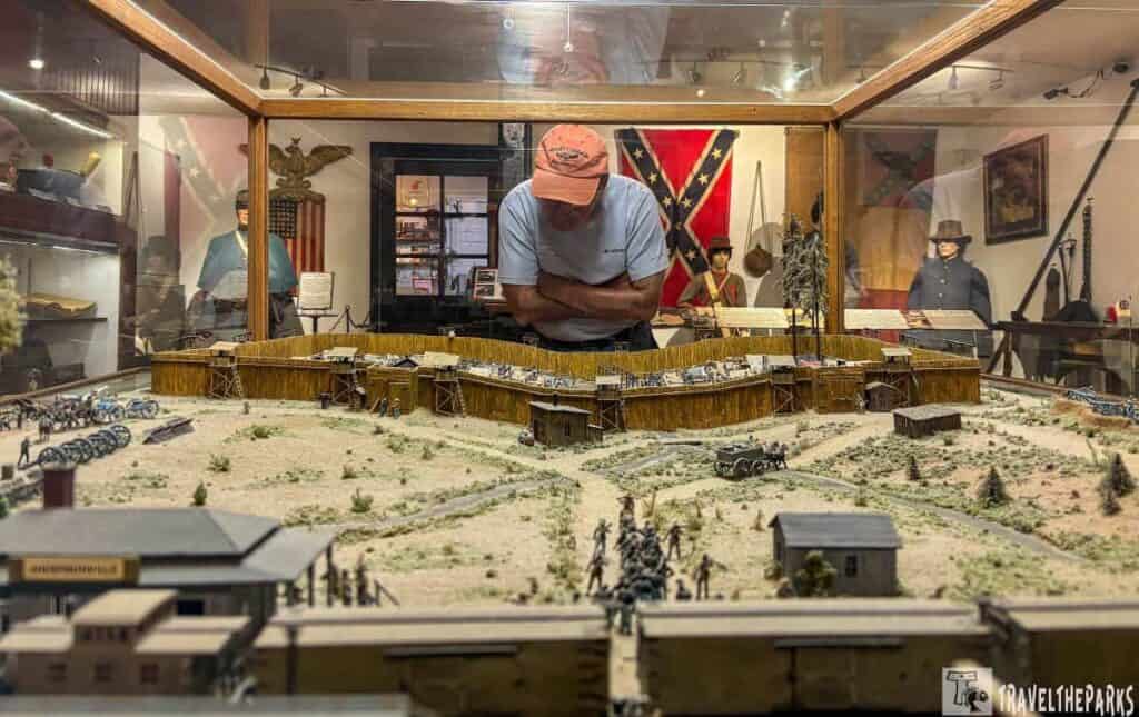 a detailed diorama displayed within a glass case, showcasing a historical scene at the Andersonville National Historic Site. The diorama features a miniature representation of a military fortification, complete with structures, cannons, and small model figures. The scene portrays a dirt and grass landscape with pathways and trees scattered throughout. In the background, a man wearing an orange cap and light blue shirt leans over the glass case to view the diorama closely. Behind him, several mannequins dressed in historical military uniforms stand against a backdrop that includes an American flag and a Confederate flag. The setting suggests a museum or exhibit room, with other artifacts and historical displays visible along the walls.