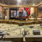 a detailed diorama displayed within a glass case, showcasing a historical scene at the Andersonville National Historic Site. The diorama features a miniature representation of a military fortification, complete with structures, cannons, and small model figures. The scene portrays a dirt and grass landscape with pathways and trees scattered throughout. In the background, a man wearing an orange cap and light blue shirt leans over the glass case to view the diorama closely. Behind him, several mannequins dressed in historical military uniforms stand against a backdrop that includes an American flag and a Confederate flag. The setting suggests a museum or exhibit room, with other artifacts and historical displays visible along the walls.