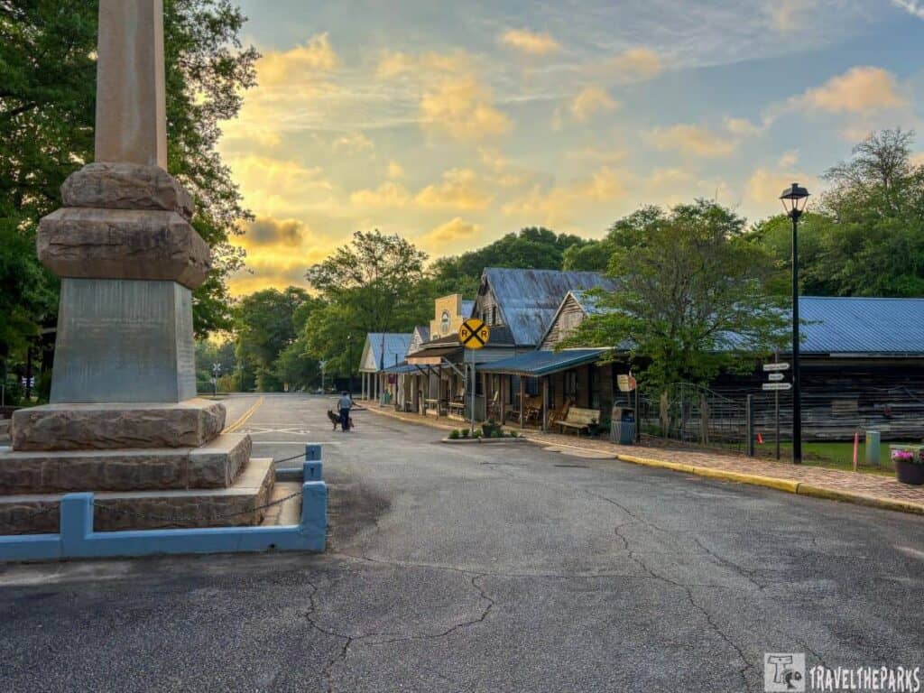 at sunset in the town of Andersonville. On the left, the
Henry Wirz Monument with a smooth column and textured base stands on a broad pedestal. The street extends into the background, flanked by quaint wooden buildings with blue metal roofs. A railroad crossing sign is visible in the mid-ground. Trees line both sides of the street, lush with green foliage. The sky is partly cloudy with hints of orange and yellow from the setting sun. A streetlamp and a few people walking are visible in the distance. The pavement has a worn, textured appearance, and the scene exudes a peaceful, historic charm.