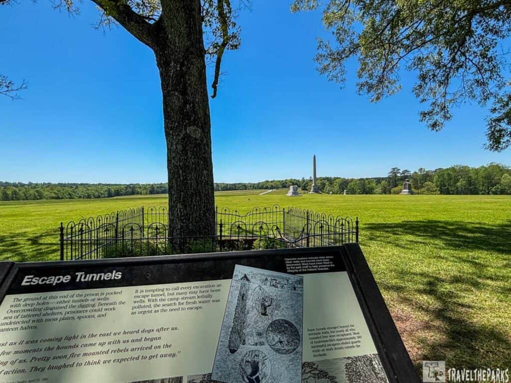 Andersonville National Historic Site. In the foreground, there is an informational plaque titled "Escape Tunnels" positioned on a stand. The plaque contains text and an illustration related to escape tunnels. A large tree with a thick trunk stands nearby, casting shade and partially framed by a small, decorative black metal fence. Further back, an expansive green field stretches towards the horizon. In the distance, several monuments, including a prominent obelisk, are visible. The sky is clear and blue, with scattered trees lining the horizon.