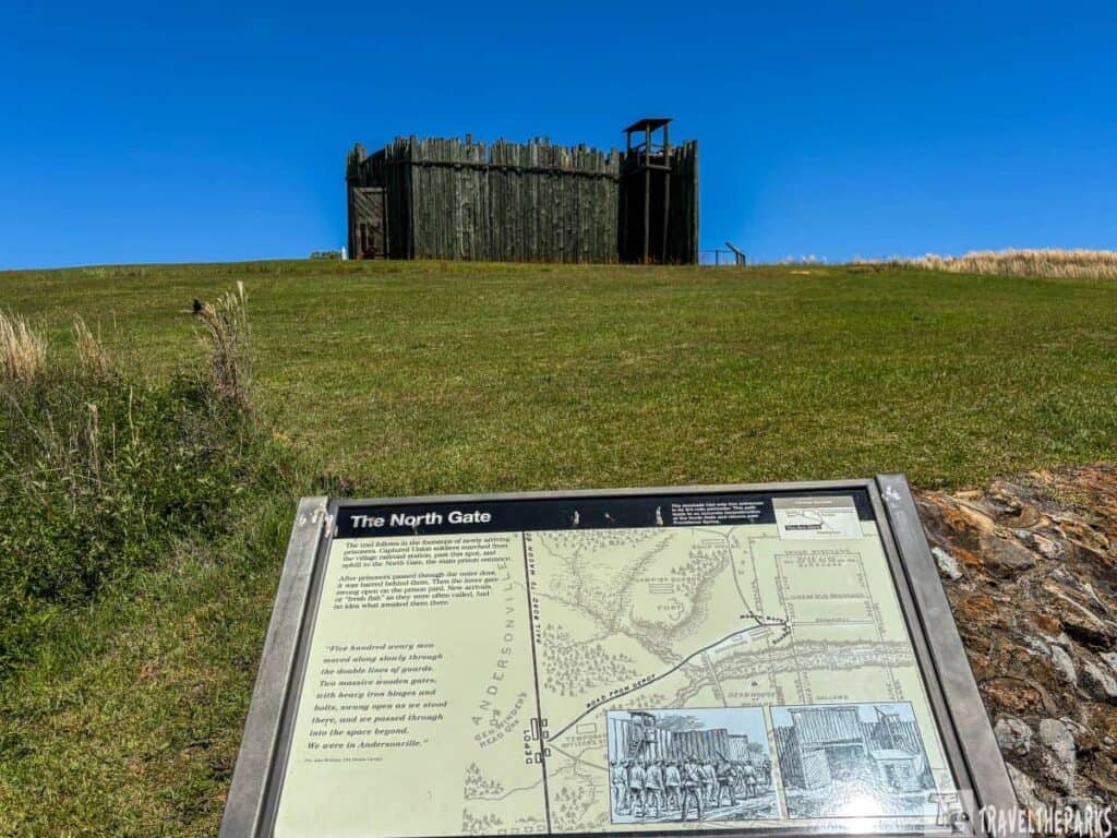 Andersonville National Historic Site. In the background, a wooden stockade structure stands on a grassy hill under a bright blue sky. The stockade is made of vertical wooden logs and features a small entrance. In the foreground, there is an interpretive sign angled toward the viewer. The sign shows a historical map and illustrations related to the site, with a focus on the structure known as "The North Gate." Surrounding the frame of the sign is short grass, while taller grasses appear to the left side.