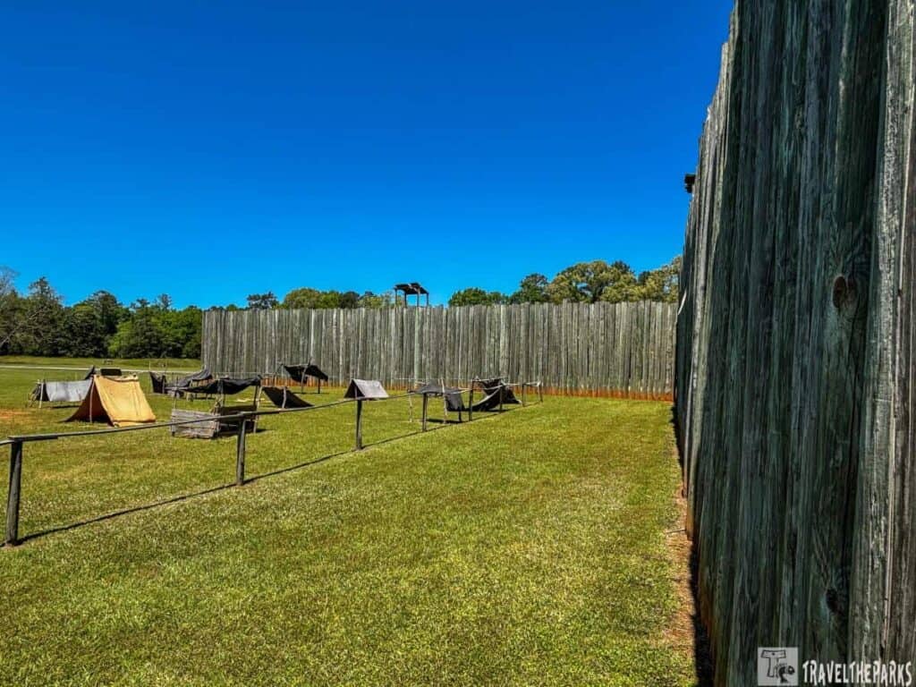 Camp Sumter Stockade, part of the Andersonville National Historic Site. The foreground features a grassy field with a row of simple wooden posts connected by rails, forming a low barrier. Within this area, several makeshift shelters, resembling small tents, are scattered across the field. These shelters are constructed from various materials, including wood and fabric, hinting at a rudimentary and temporary nature. In the background, a tall stockade wall made of vertically aligned wooden logs encircles the area. The wall extends to the horizon on the right, creating a robust enclosed environment. The sky above is clear and deep blue, suggesting a bright and sunny day.