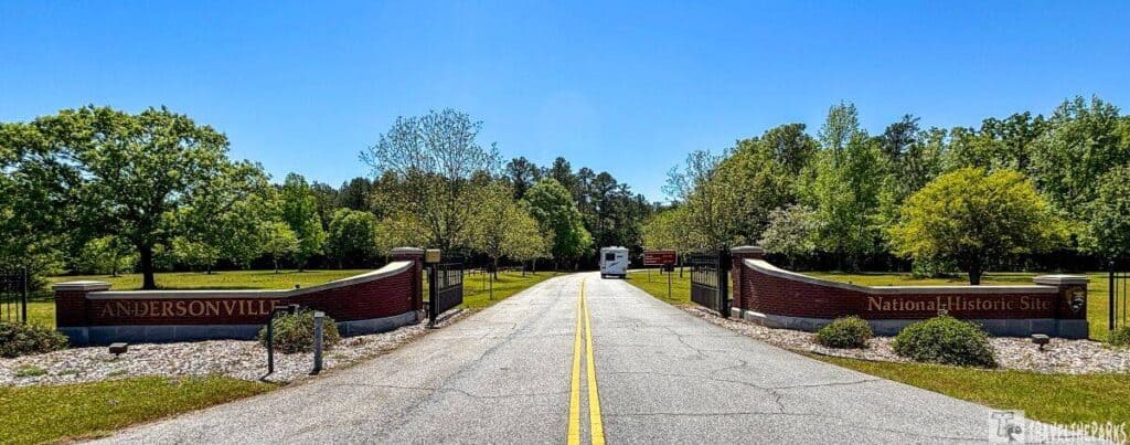 the entrance to the Andersonville National Historic Site. A straight road with dual yellow lines runs down the center, leading directly to a recreational vehicle parked further down. The road is flanked by red brick walls with iron gates on either side, conveying a sense of formality and welcome. The left brick wall bears the inscription "Andersonville," while the right states "National Historic Site." Both walls are complemented by landscaped lawns and beds of white rocks with scattered greenery, providing a neat and organized appearance. Surrounding the entrance and stretching into the background is a lush copse of trees, their leaves vibrant green under a bright blue sky, indicating a clear, sunny day.