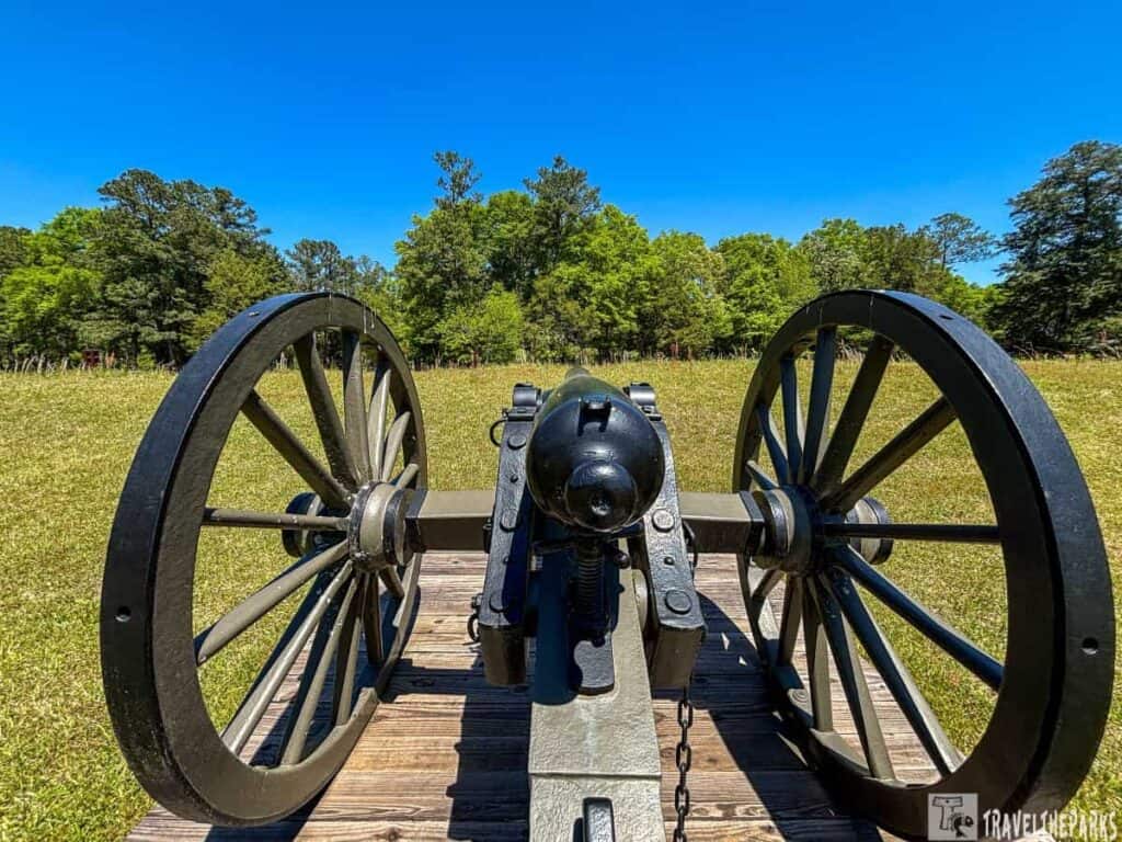 a historical cannon positioned on a grassy field under a clear blue sky. The cannon is mounted on wooden wheels with black metal bands and lies on a wooden platform. It is seen from the back, with the barrel pointing towards a distant line of lush green trees. The grass is well-maintained, and the trees vary in size and density. The sky is bright and vivid, emphasizing a sunny day.