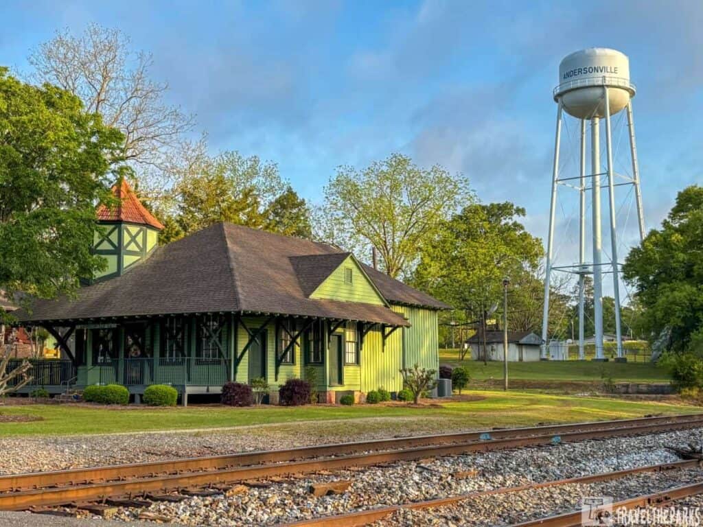a historic train depot building and a tall water tower in Andersonville. The depot, positioned on the left, is a quaint, light green structure with dark green trim and a steep, shingled roof. It features a small tower with a reddish-brown roof on the left side. In front of the building is a well-maintained lawn with small shrubs and a tree. In the foreground are railway tracks stretching horizontally across the image. On the right, the water tower stands tall against a blue sky with slight cloud coverage. The landscape is lush with a mix of large and small trees, indicating a late afternoon with warm lighting