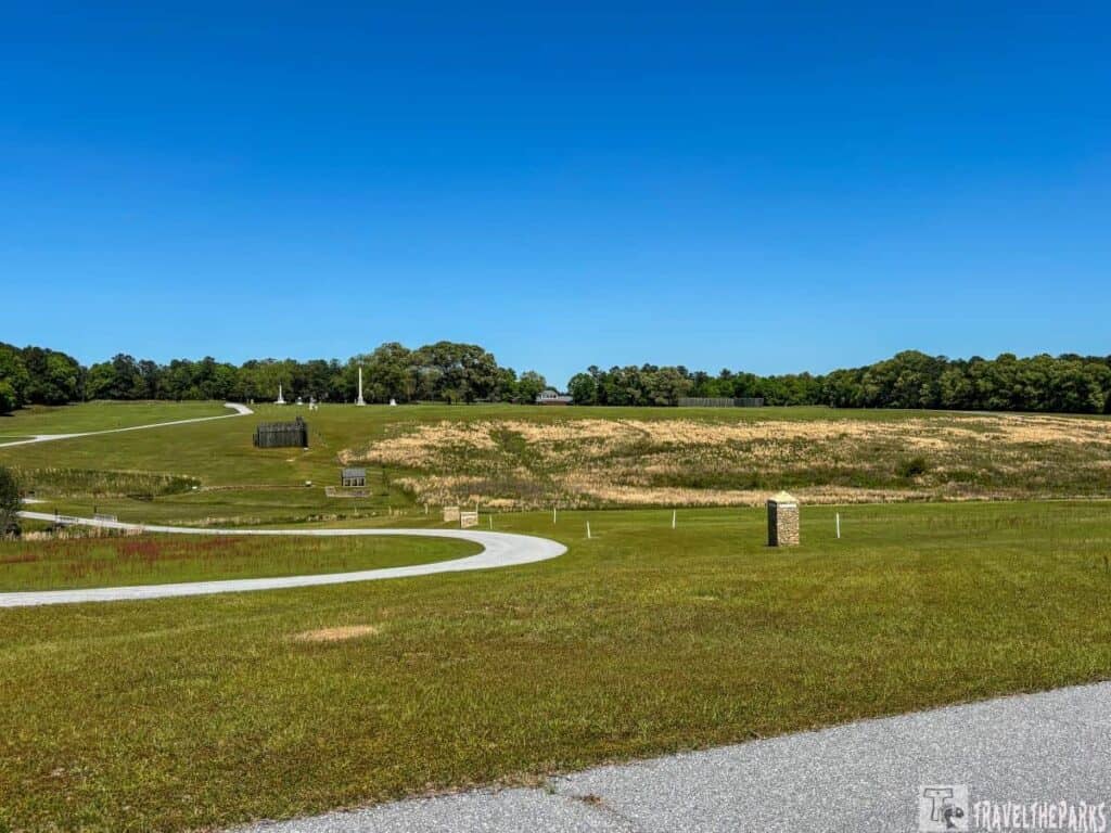 Andersonville National Historic Site. In the foreground, there is a paved walkway curving through a grassy area. The midground features a small, dark wooden structure and several stone monuments. Behind these, fields of tall, pale grasses stretch towards a line of dense trees in the background. The sky is clear and vividly blue, providing a stark contrast to the greenery and earth tones below.
