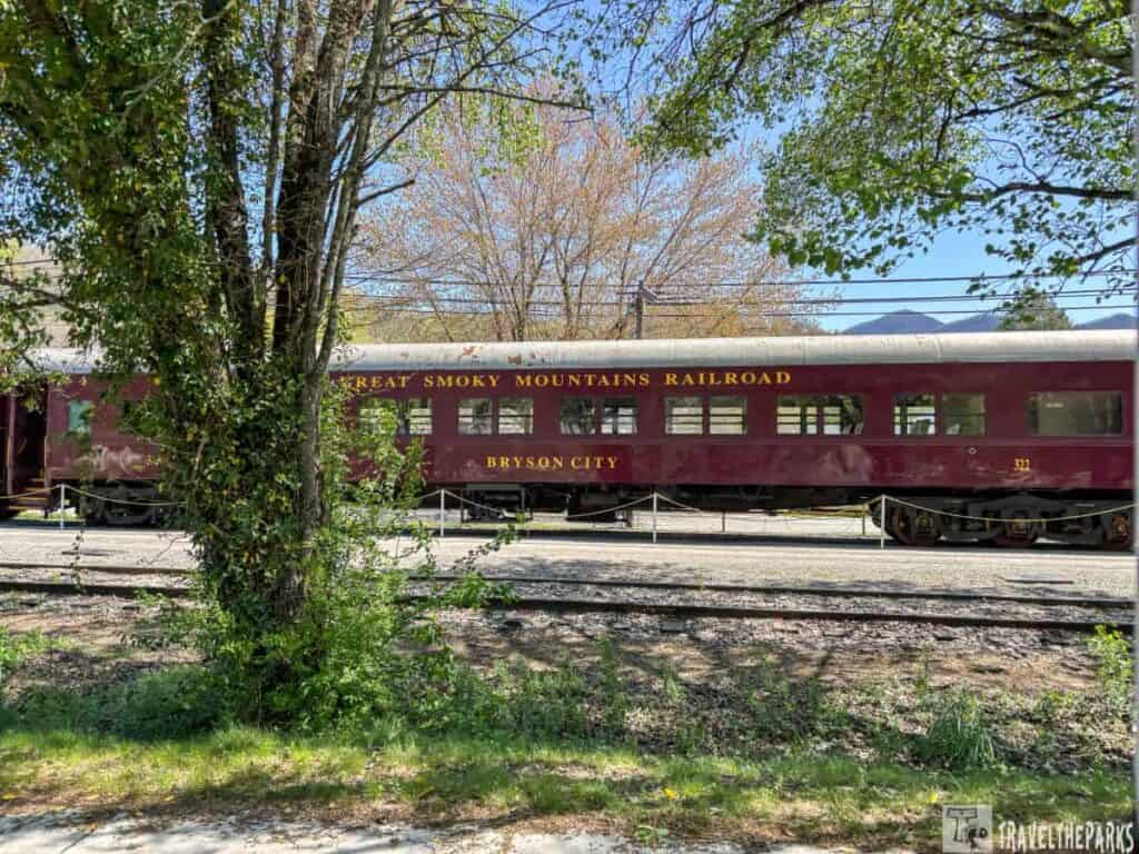 Burgundy train car labeled "Great Smoky Mountains Railroad" and "Bryson City" on tracks, with trees and a mountain in the background.