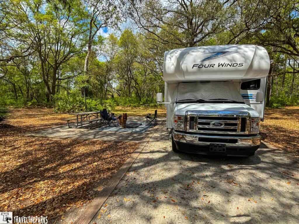 a campsite at Reed Bingham State Park, featuring a parked "Four Winds" RV on a paved surface surrounded by a natural forest setting. The RV is positioned on the right side, with its front facing the viewer. Its white exterior contrasts with the surrounding greenery. The windshield has a reflective sunshade. To the left, there is an empty picnic table and two foldable chairs. A dog stands near the chairs on a rug, which is partially visible. The ground is scattered with dry leaves, and tall trees with abundant green foliage frame the background, filtering sunlight and creating dappled shadow patterns on the ground.