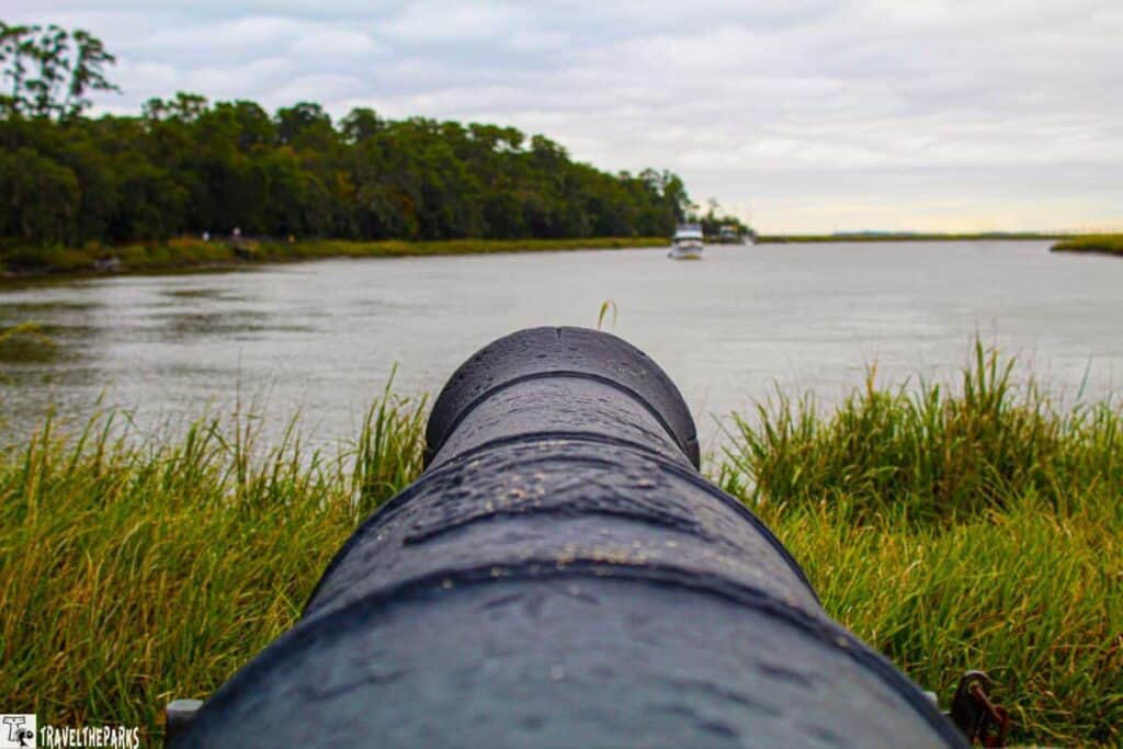 Cannon facing a river with a boat in the distance, bordered by trees under a cloudy sky.