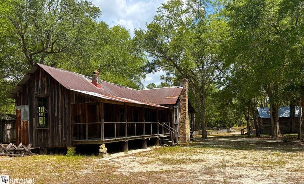A rustic wooden Cracker cabin with a rusty metal roof and brick chimney in a wooded area.