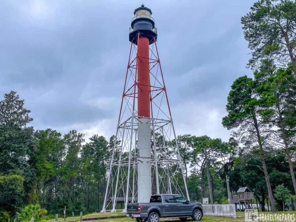 Crooked River Lighthouse in Carrabelle, Florida. The lighthouse is tall and slender, with a red and white color scheme. It consists of a white, tapered lower section supported by a lattice of metal beams, transitioning to a red upper section beneath the lantern room, which is painted black. The lighthouse is surrounded by a dense growth of green trees, suggesting a forested area. In front of the lighthouse, there is a black pickup truck parked on a grassy area.